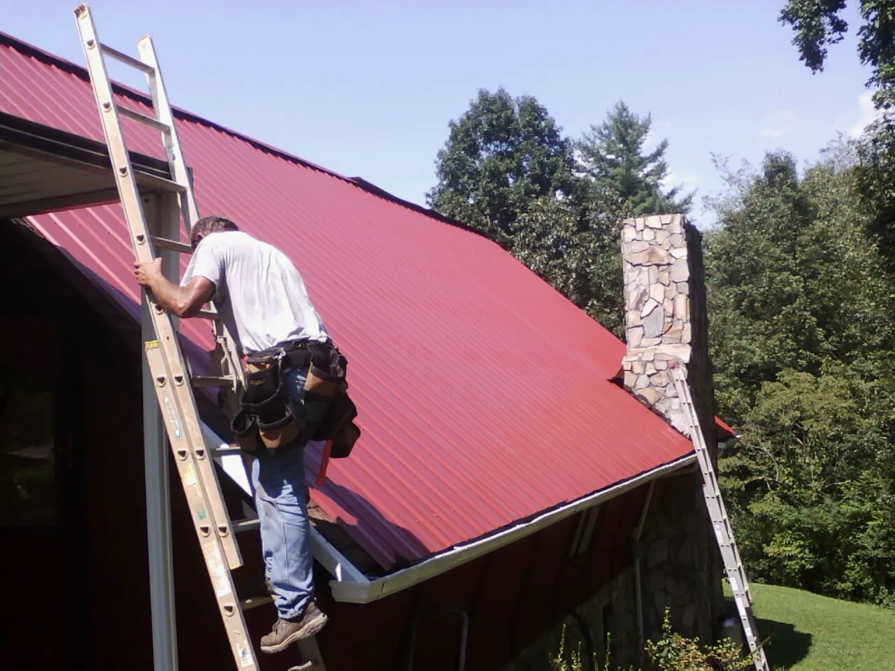 Roofer on a ladder beside a red metal roof and stone chimney — Soffit & Fascia Repair by Roofing Brothers in Hazel Park